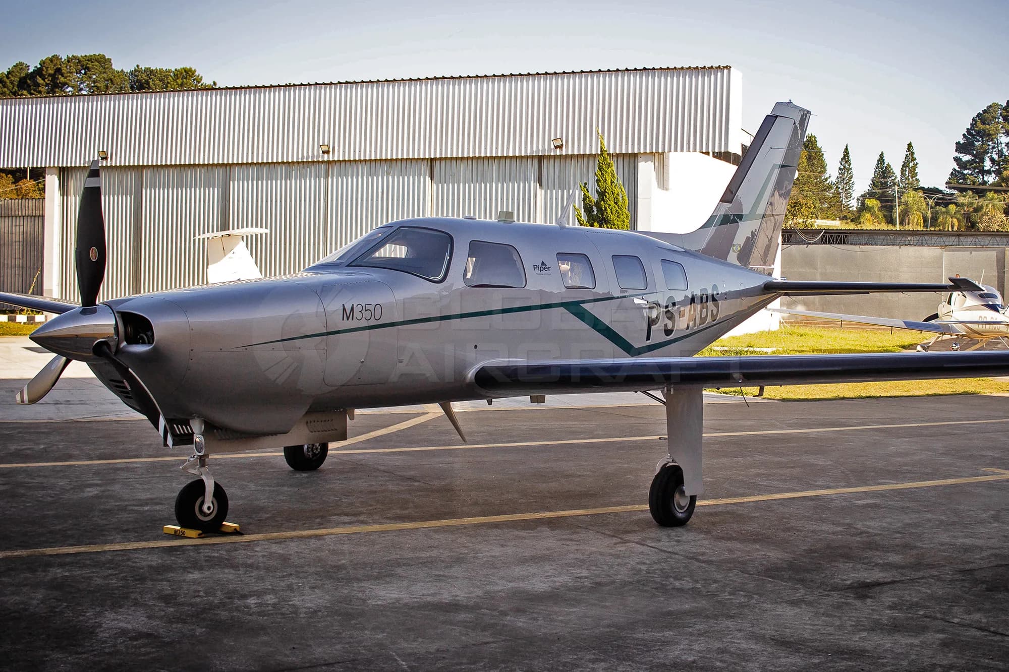 A silver Piper M350 aircraft with green and white stripes, tail number PS-ABS, parked on an tarmac with hangars in the background.