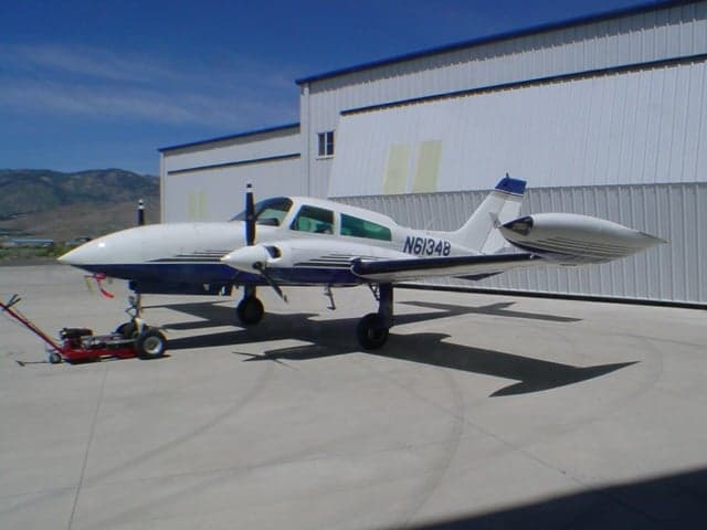 A white and blue twin-engine Cessna 310R aircraft, registration N61348, is parked on a concrete ramp next to a white hangar.