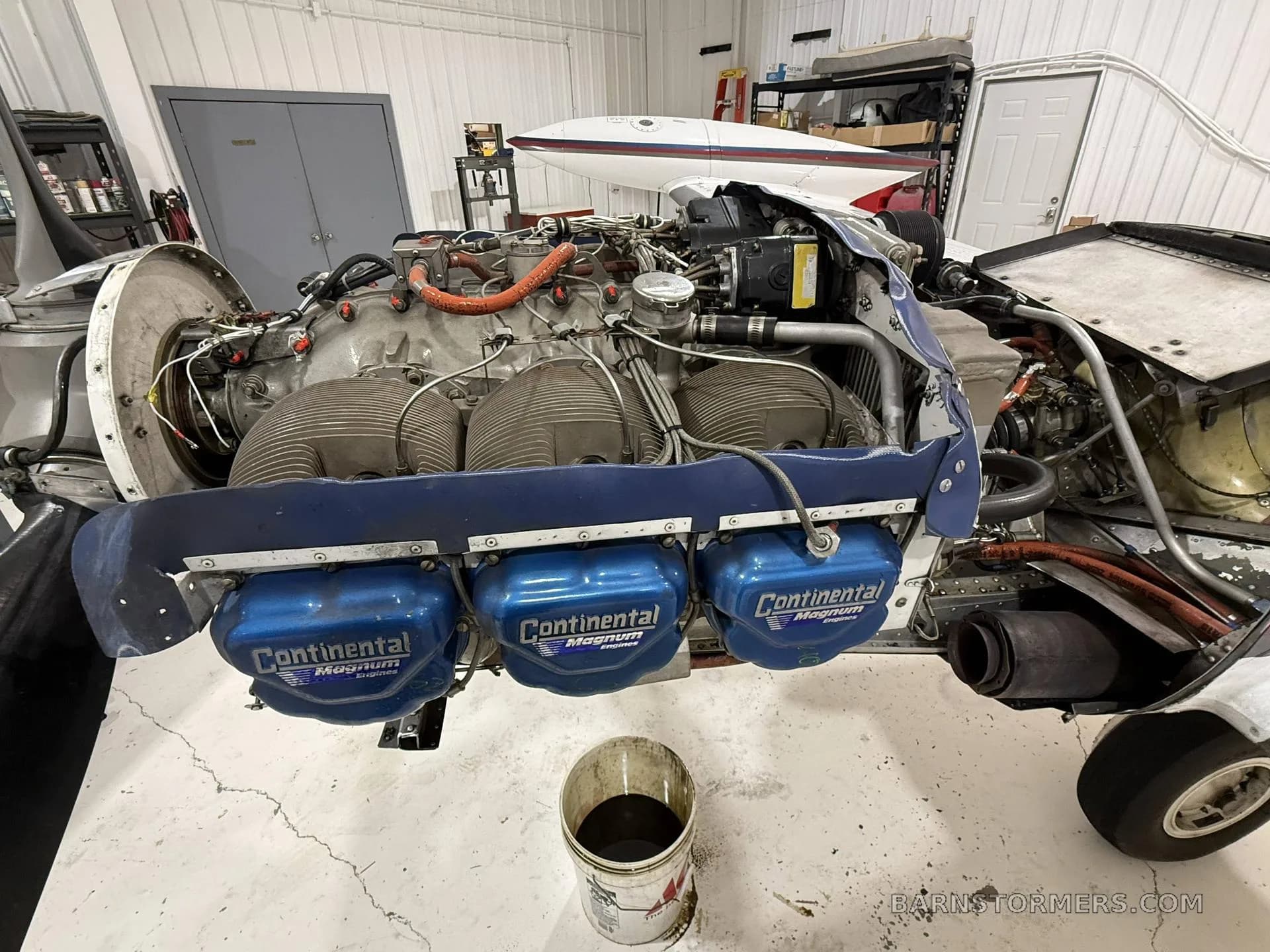 A close-up view of a Continental aircraft engine with blue valve covers exposed for maintenance, with a bucket of oil underneath, in a hangar.