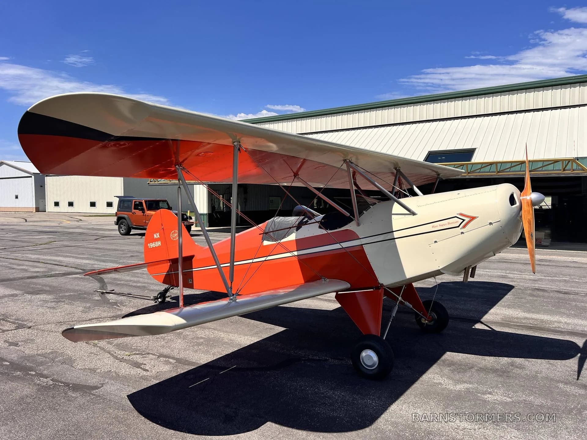 A red and white biplane with a wooden propeller is parked on an asphalt tarmac next to a hangar.
