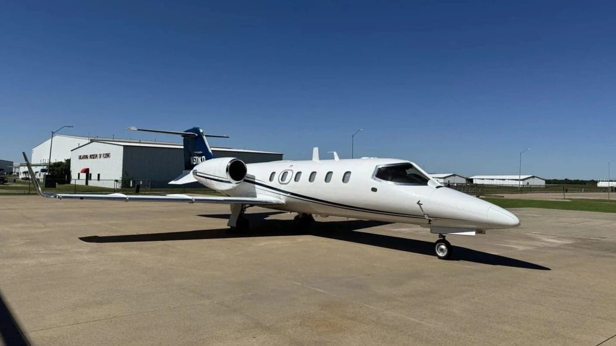 A white Learjet 31A with blue and black stripes on the tail and fuselage, parked on an airport tarmac under a clear blue sky.
