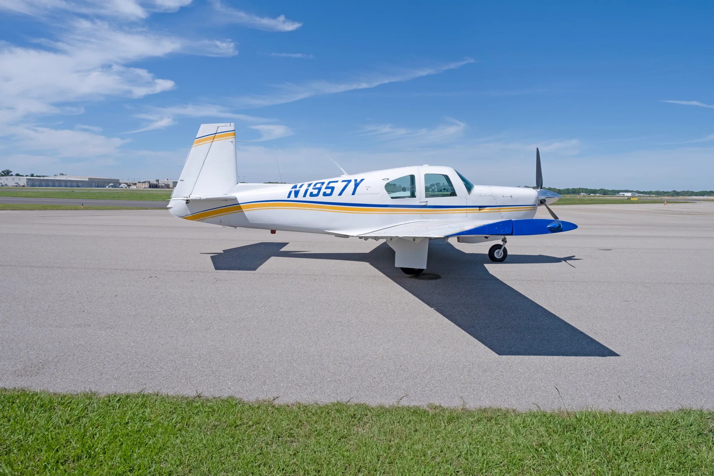 A side view of a white single-engine propeller aircraft with blue and yellow stripes, registration N1957Y, parked on an asphalt apron.