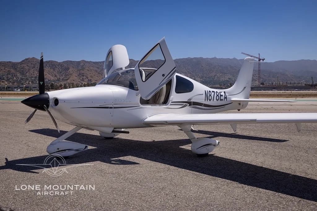 A white Cirrus aircraft with registration N878EA and open doors is parked on an airfield with mountains in the background.