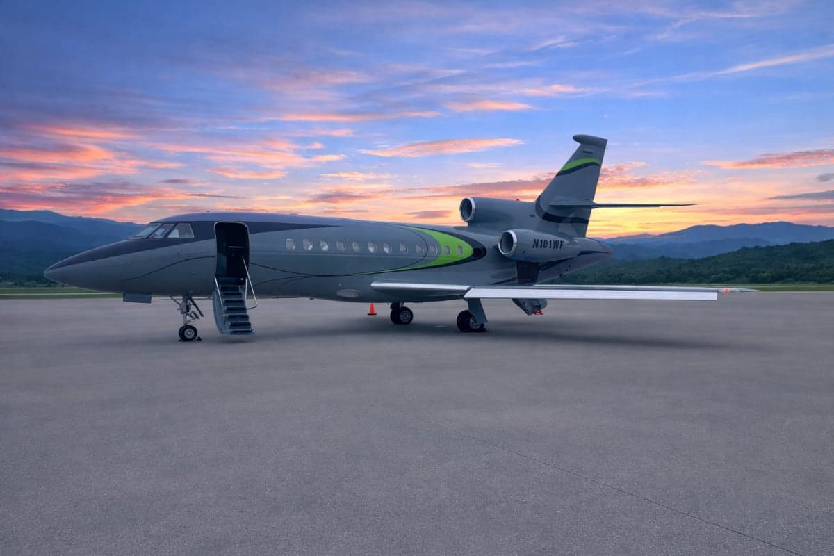A grey and green Dassault Falcon 900EX business jet with its door open and stairs extended, parked on a tarmac at sunset with mountains in the background.