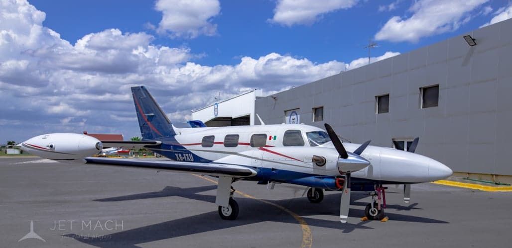 A white and blue twin-engine propeller aircraft, registration XB-FXU, is parked on a tarmac next to a white building.