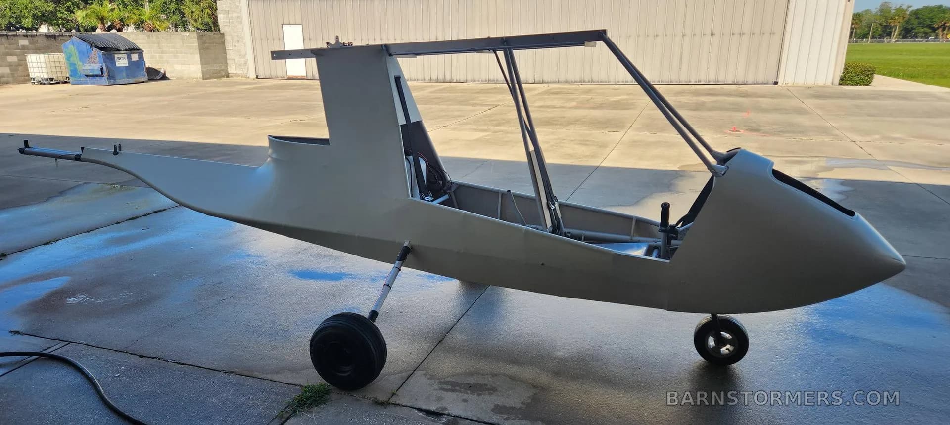 A light grey aircraft fuselage with landing gear and partial frame structure, sitting on a wet concrete pad outside a hangar.