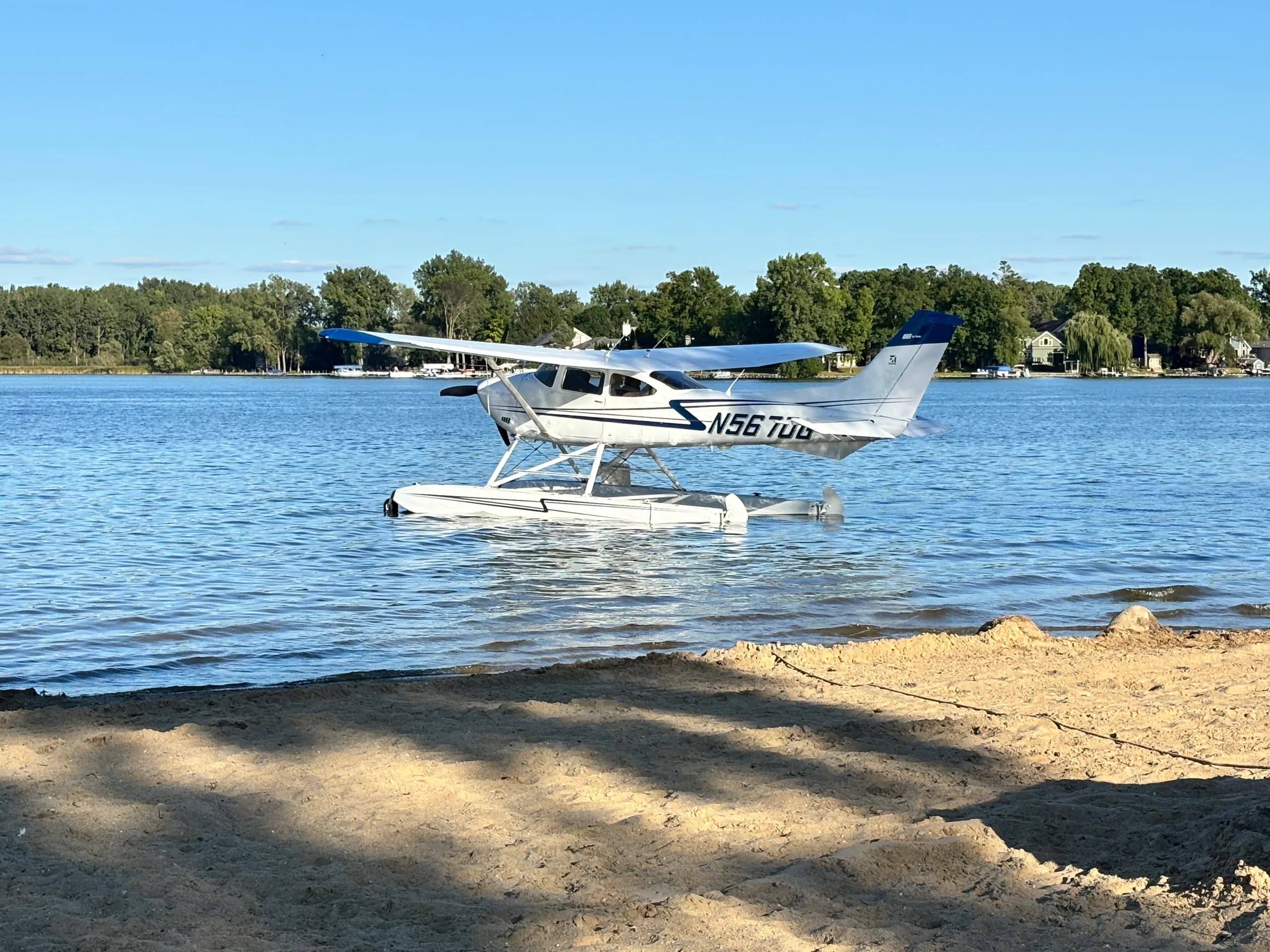 A white floatplane, N567DG, with blue accents, resting on the water near a sandy shore.