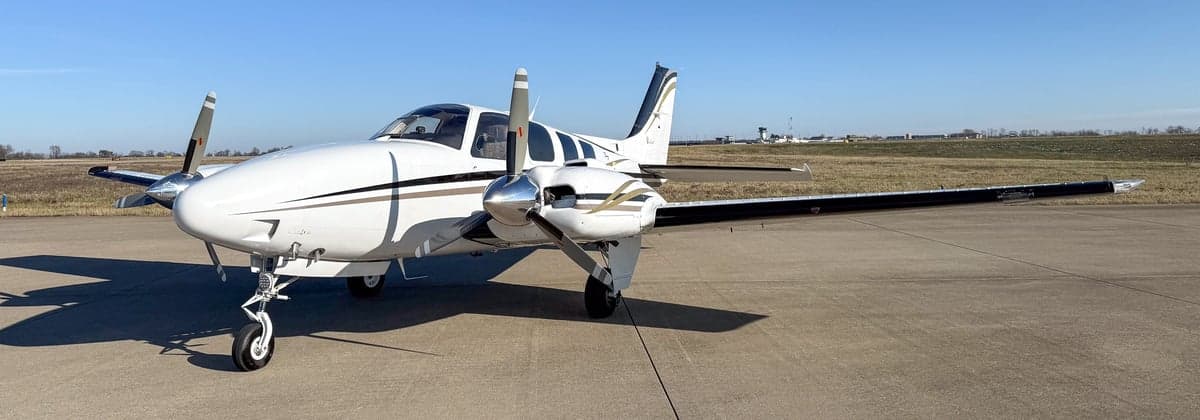 Front-right view of a white twin-engine propeller aircraft with black and gold stripes on an airfield.