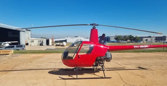 A red ZU-180 helicopter is parked on an airfield tarmac with a person working near its engine.