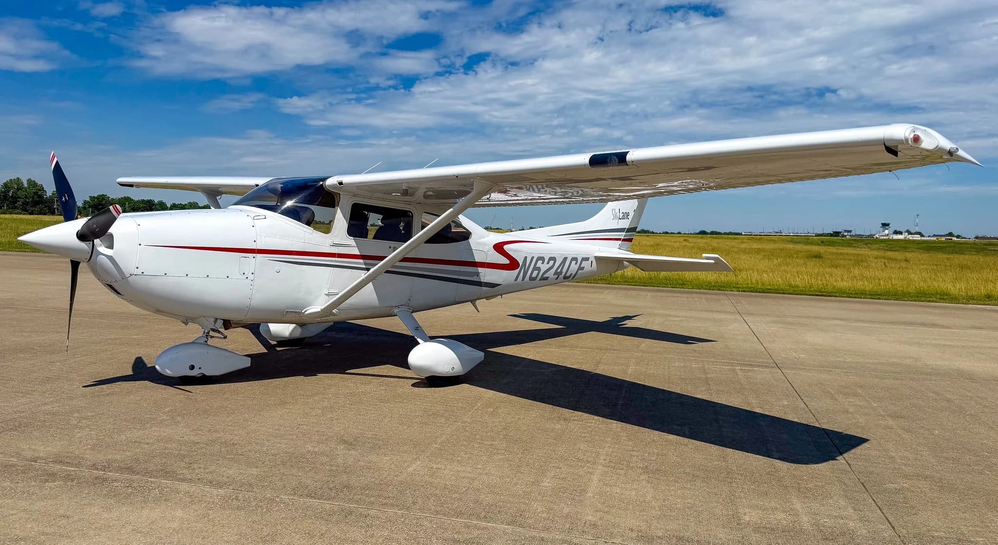 A white Cessna 182 Skylane with red and gray stripes, registration N624CF, parked on an asphalt ramp under a partly cloudy blue sky.