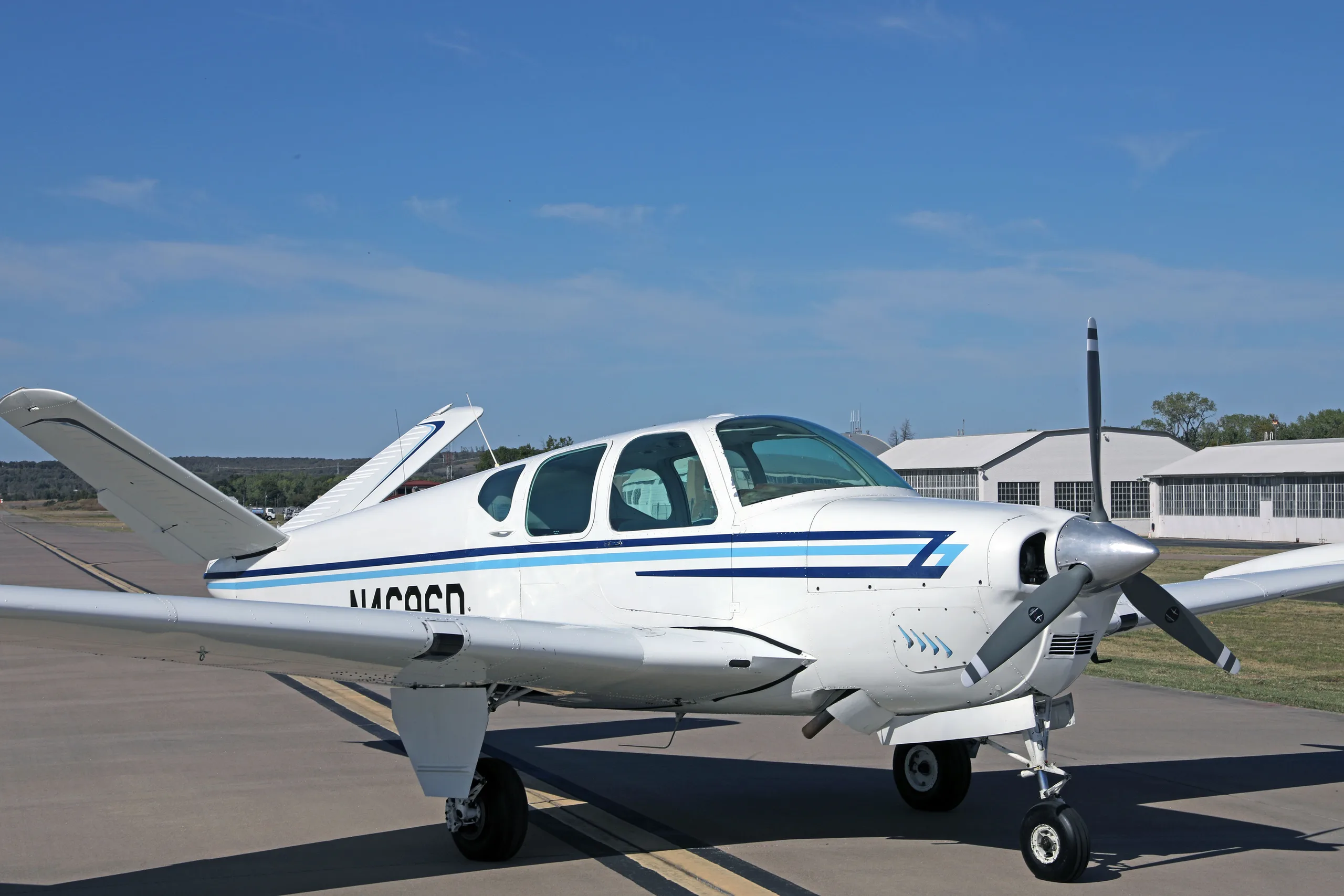 A white single-engine aircraft with blue stripes, registration N4696D, parked on an airfield with hangars in the background.
