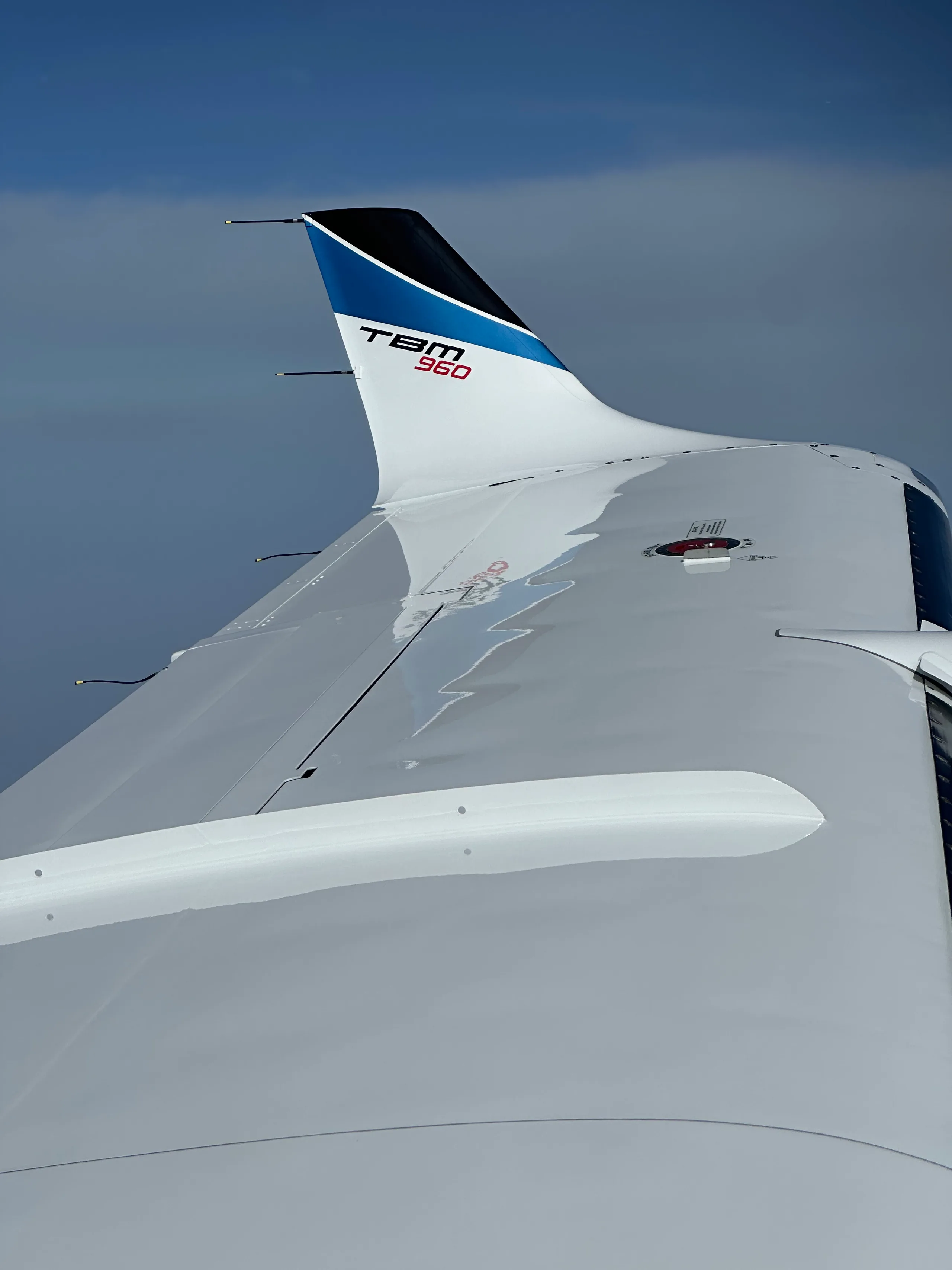 An aerial view of the white wing and blue-tipped tail of a TBM 960 aircraft against a blue sky.