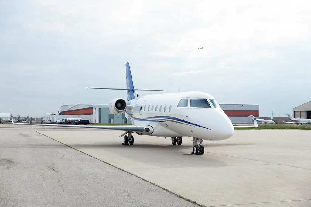 A white business jet with blue stripes and tail, parked on an airport tarmac with other aircraft and hangars in the background.
