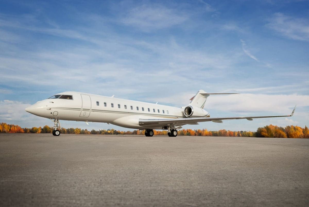 A white jet sits on the tarmac with fall foliage in the background.