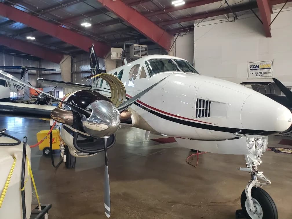 A white Beechcraft King Air 90 turboprop aircraft with black and red stripes on the fuselage, parked in a hangar, viewed from the front-right.