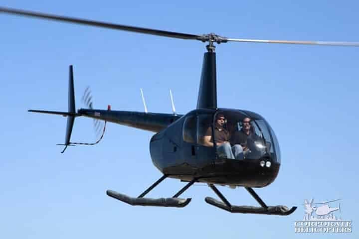 A black Robinson R44 helicopter with two people inside, flying against a clear blue sky.