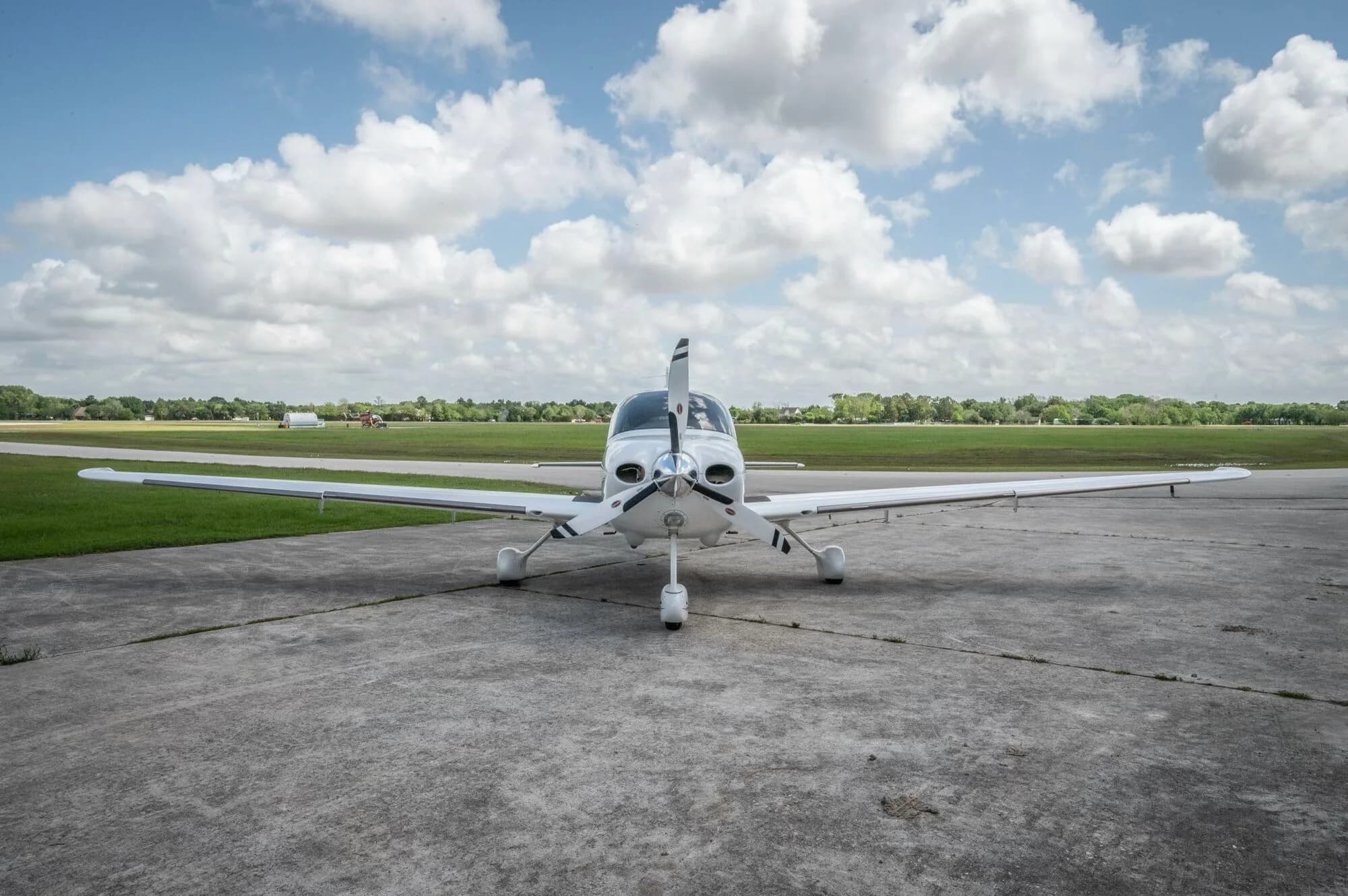 A head-on view of a white Cirrus SR22 GTS aircraft with a three-bladed propeller parked on a concrete ramp.