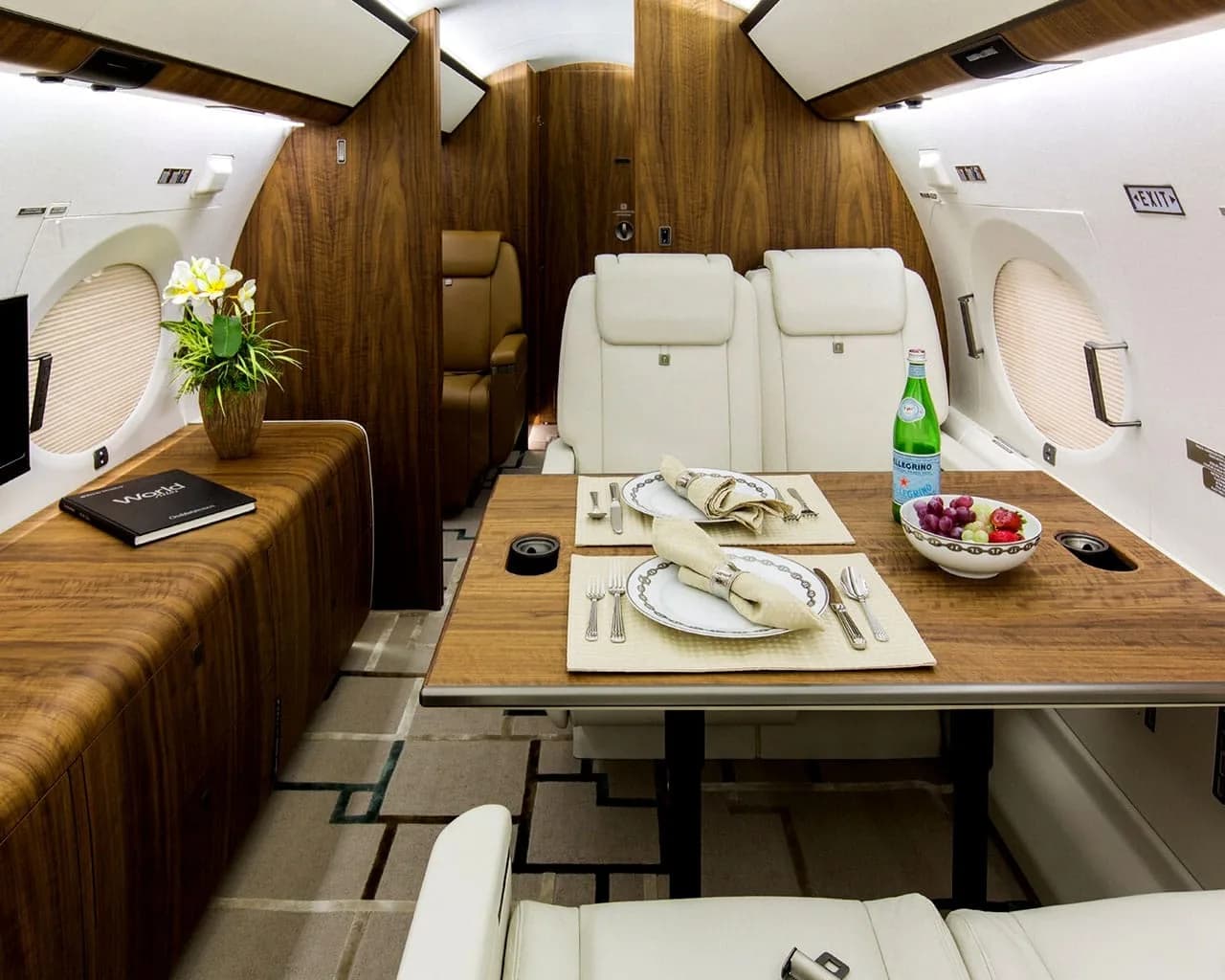 A private jet cabin dining area with a set table for two, white leather seats, and dark wood accents.