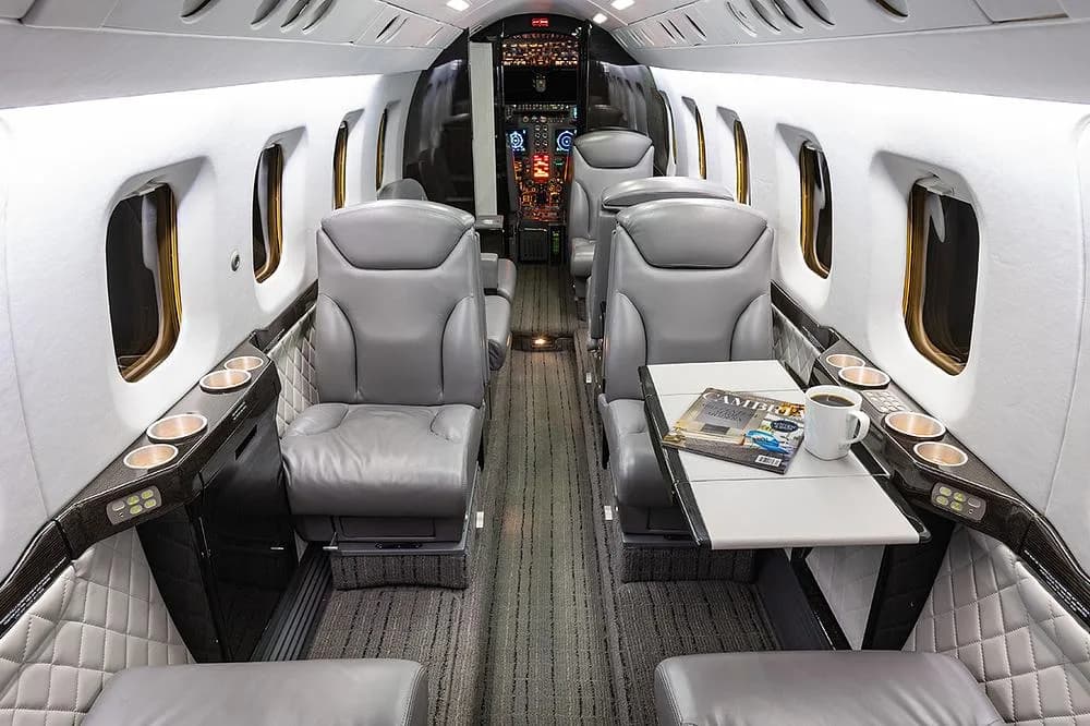 The luxurious gray leather interior of a business jet cabin with a table, magazines, and a coffee cup.