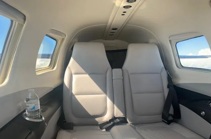 The aircraft's cabin interior shows two light grey seats and windows with a view of clouds.