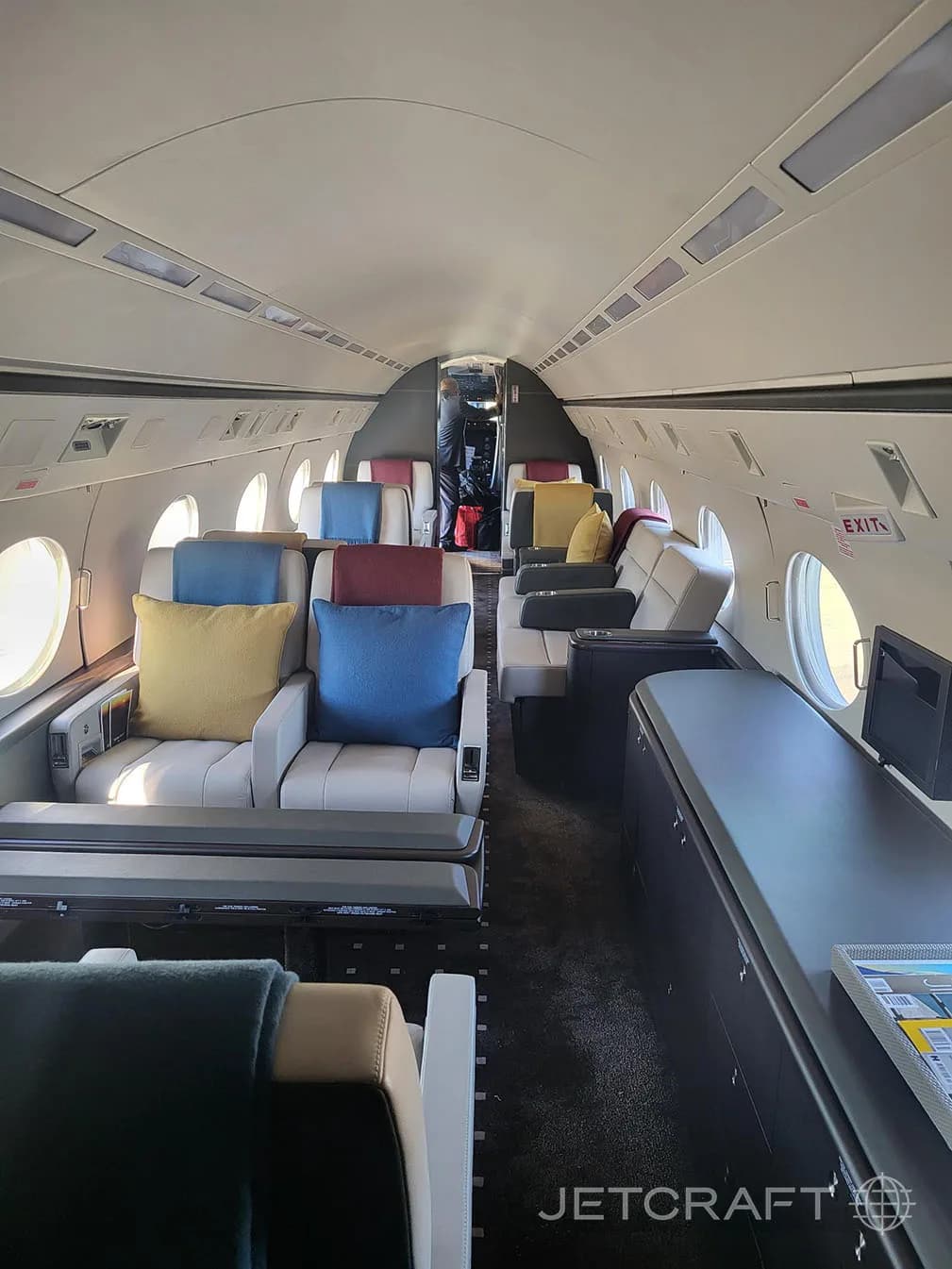 The forward section of a business jet cabin with light grey seats, colorful pillows, and a dark credenza on the right.
