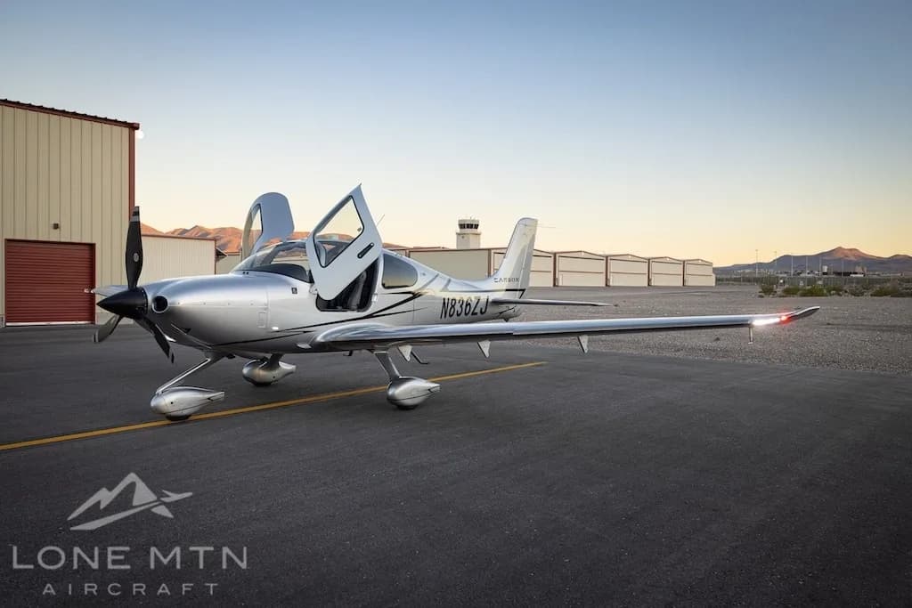 A silver Cirrus SR22T aircraft with registration N836ZJ is parked on an asphalt ramp with its doors open, facing a control tower and hangars at sunset.
