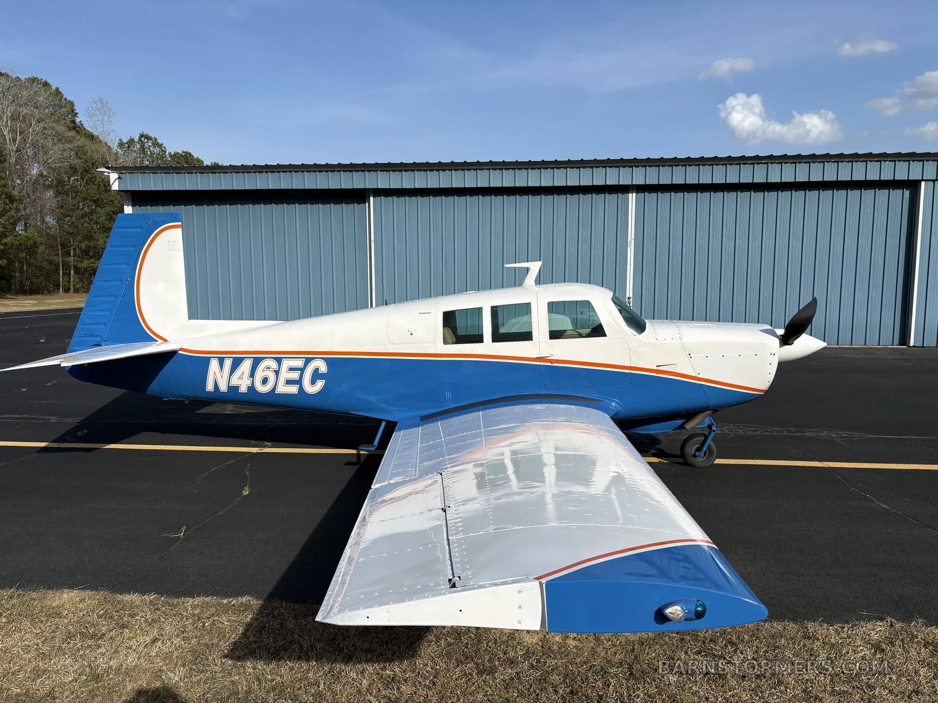 A white and blue single-engine aircraft with tail number N46EC parked on an asphalt ramp next to a blue hangar.
