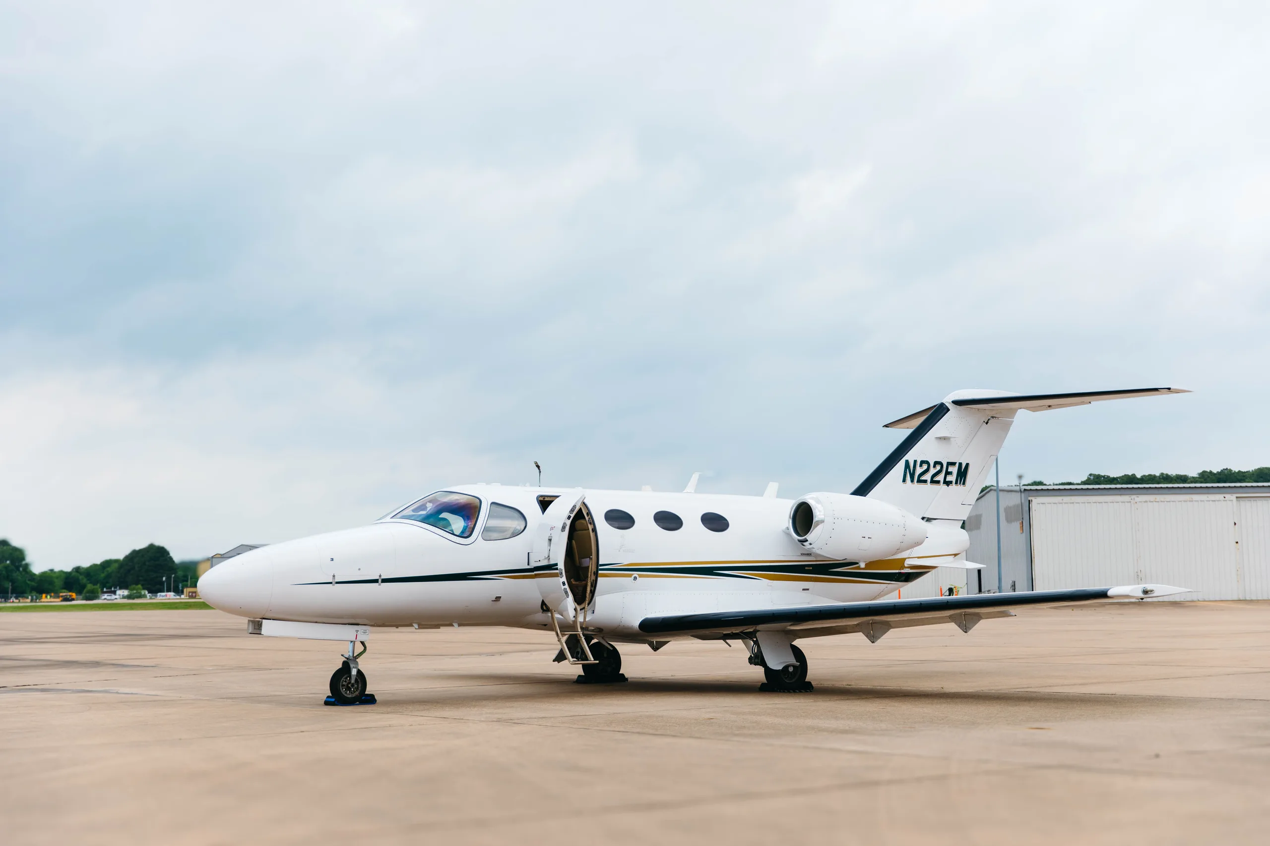 A white Cessna Citation M2 business jet, tail number N22EM, with its main cabin door open, parked on a tarmac with hangars in the background.