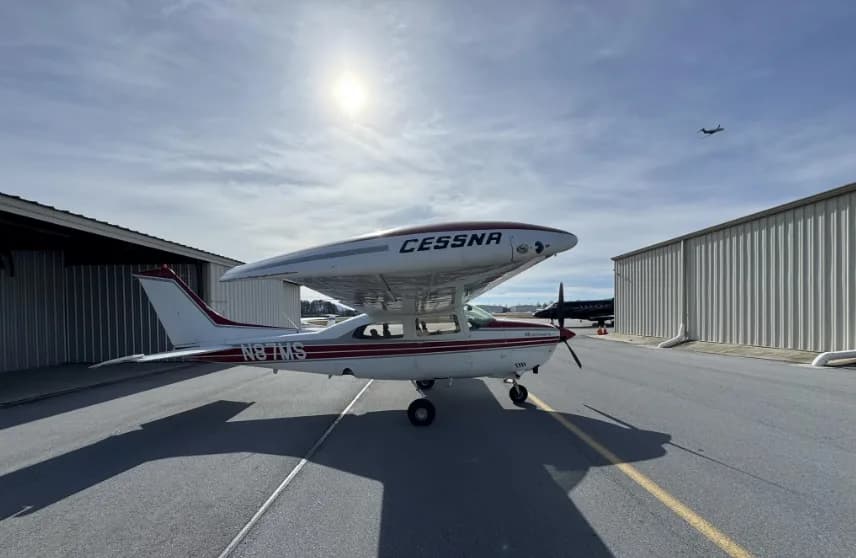 A front-right view of a white and red Cessna 182 Skylane (N87MS) parked on an airport tarmac under a bright sky.