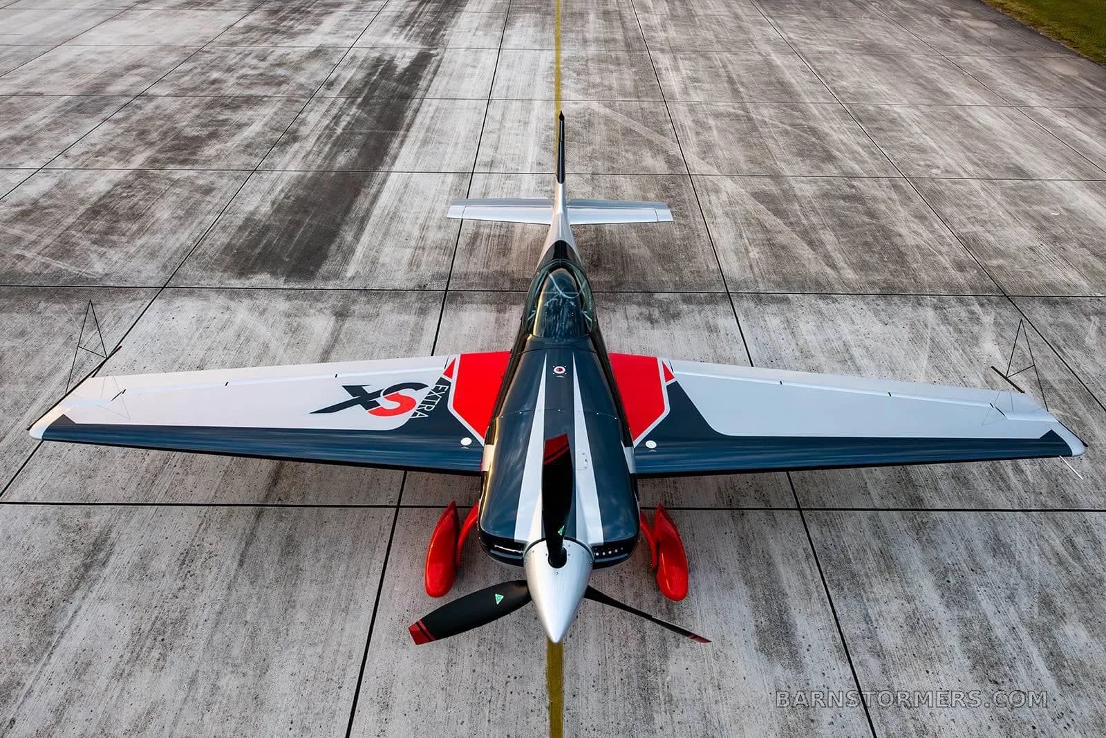An overhead front view of a red, black, and silver Extra 330SC aerobatic aircraft parked on a concrete runway with a yellow centerline.