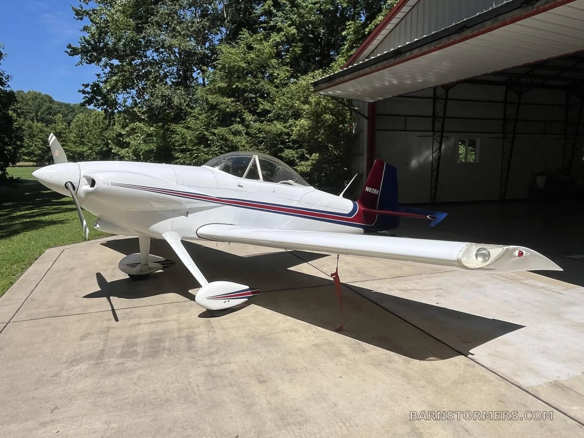 A white single-engine aircraft with red and blue stripes on the tail and fuselage, parked on concrete in front of an open hangar with trees in the background.