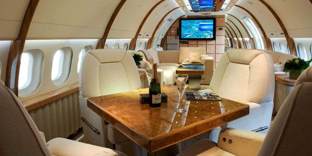 A luxurious private jet interior features cream-colored seats, wood paneling, and a bottle of champagne.