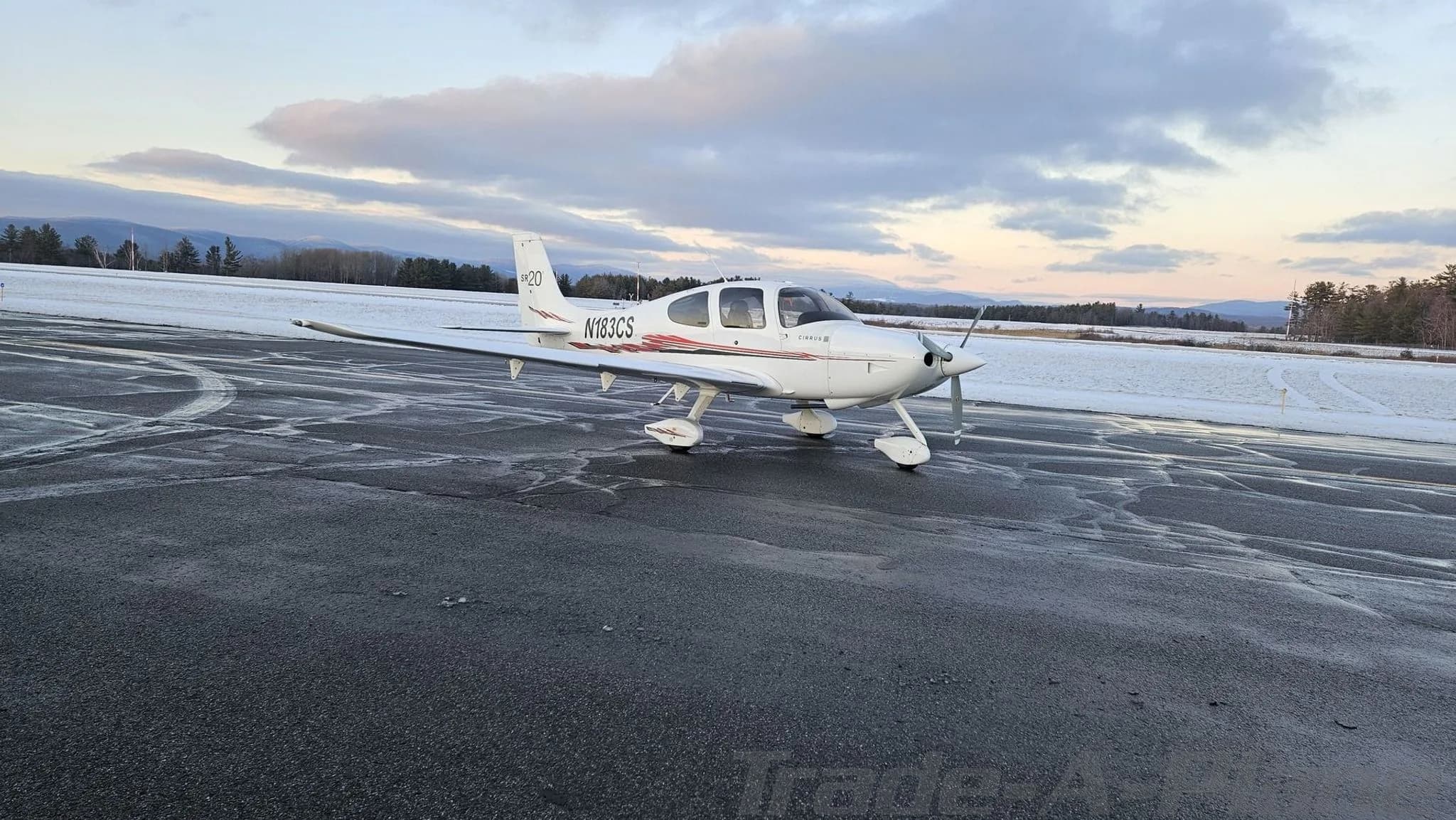 A white Cirrus SR20 aircraft with red and black stripes on a wet tarmac at sunset.