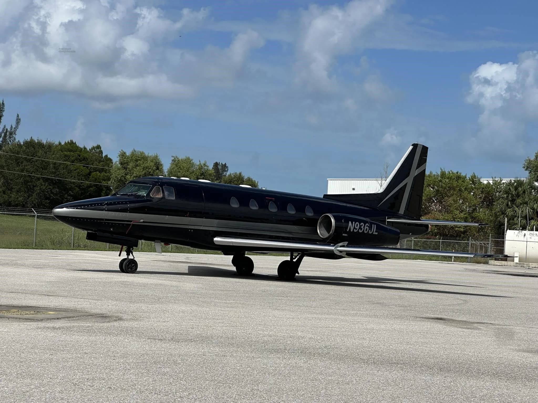 A black and silver business jet, N936JL, parked on an asphalt tarmac with trees and buildings in the background.