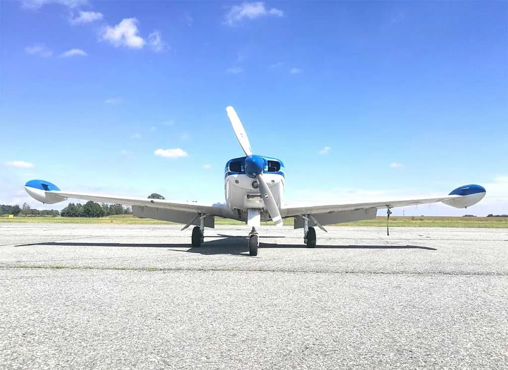 A white and blue single-engine aircraft with wingtip tanks is viewed head-on from a low angle on an asphalt runway under a clear blue sky.