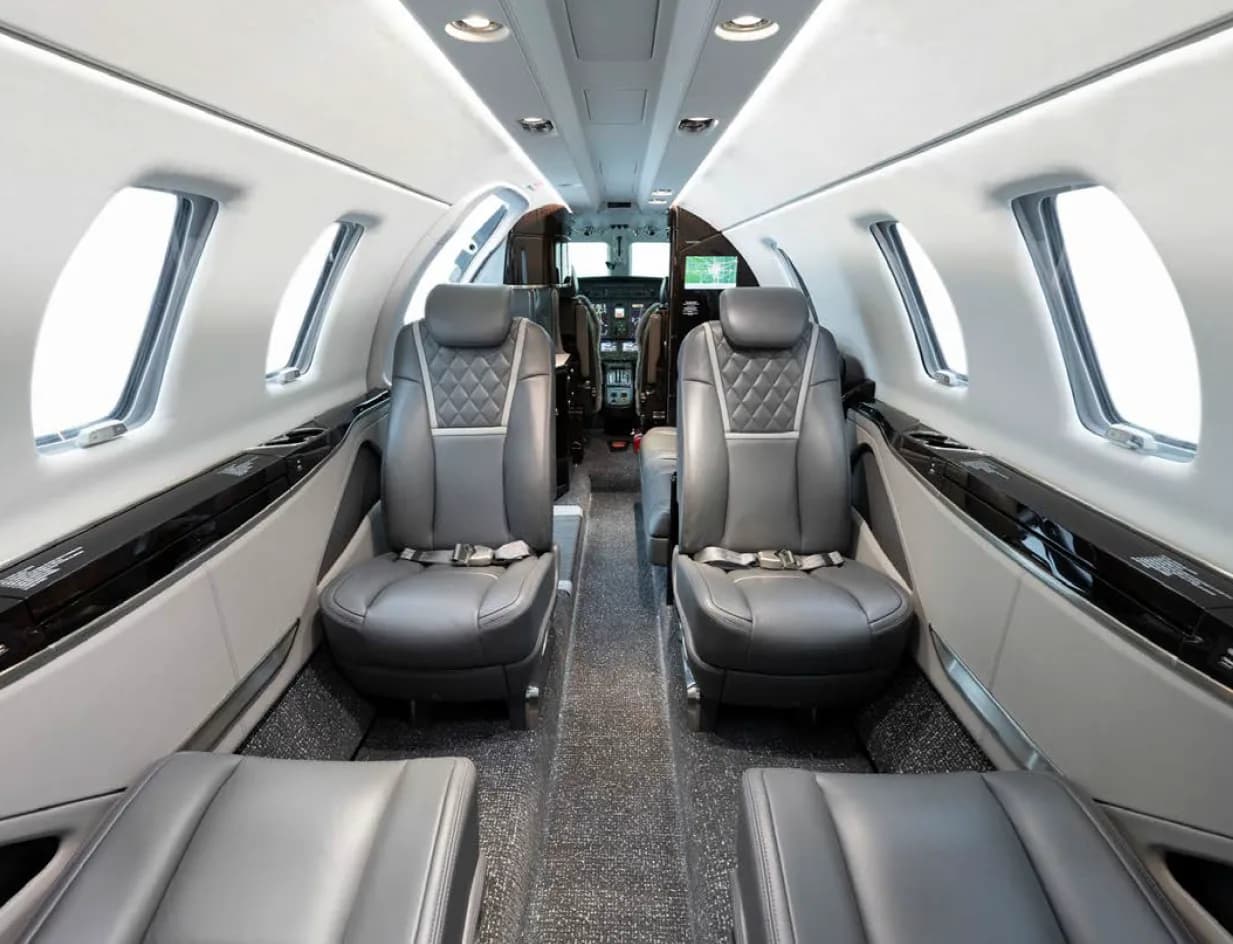 A view down the aisle of a business jet cabin, showing two rows of grey leather seats and the cockpit in the distance.