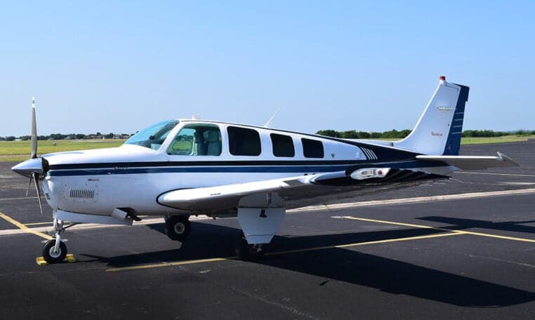 A white and blue Beechcraft Baron aircraft is parked on an asphalt tarmac under a clear sky.