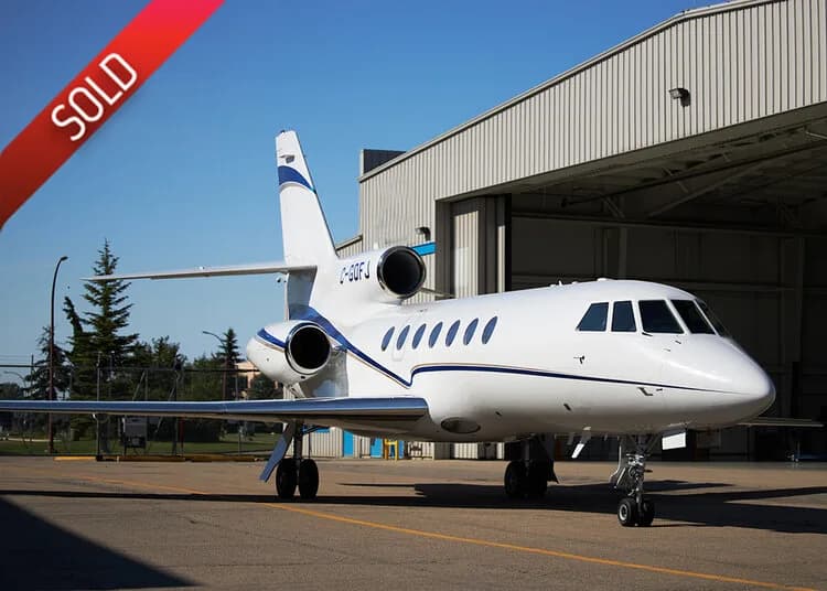 A white Dassault Falcon 900EX business jet with blue accents is parked on a tarmac next to a hangar under a clear sky.