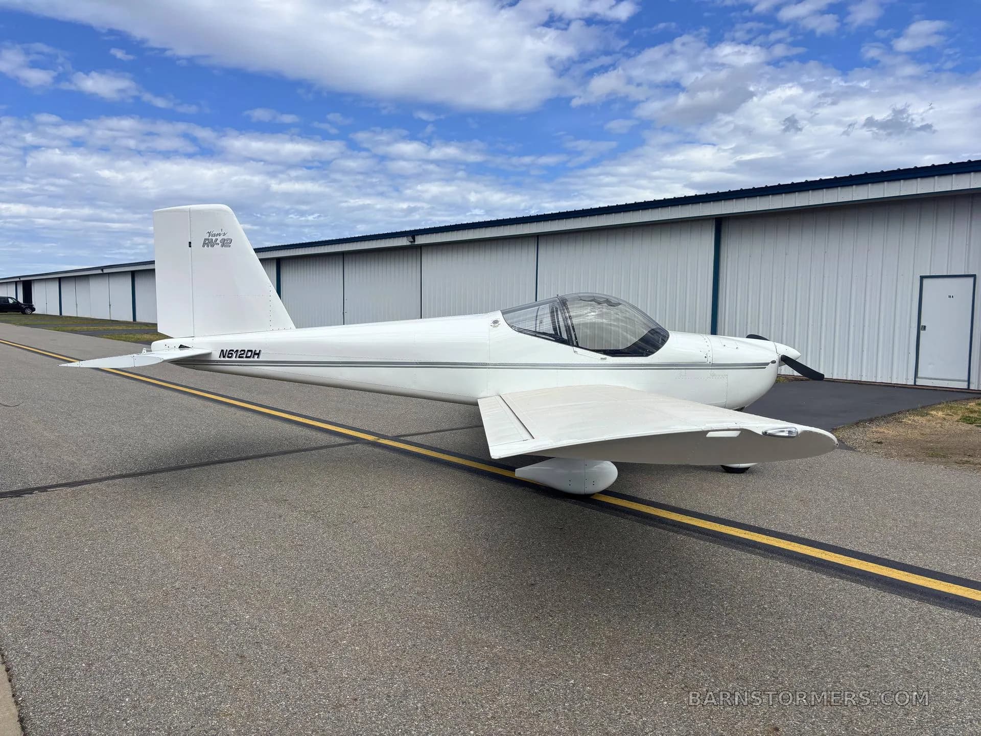 A white Vans RV-12 aircraft (N612DH) is parked on an airfield tarmac next to a long white hangar under a partly cloudy sky.