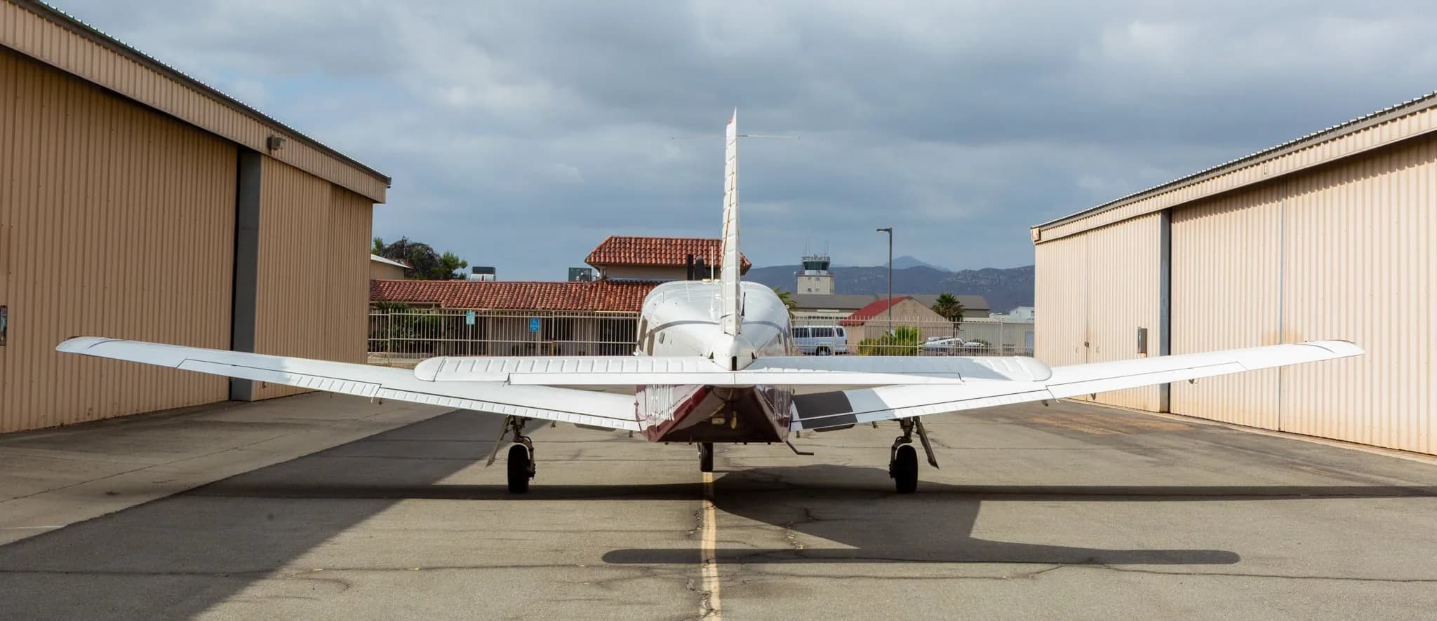 Rear view of a white and maroon Piper Saratoga II TC aircraft, registration N691MA, parked between hangars with a control tower in the distance.