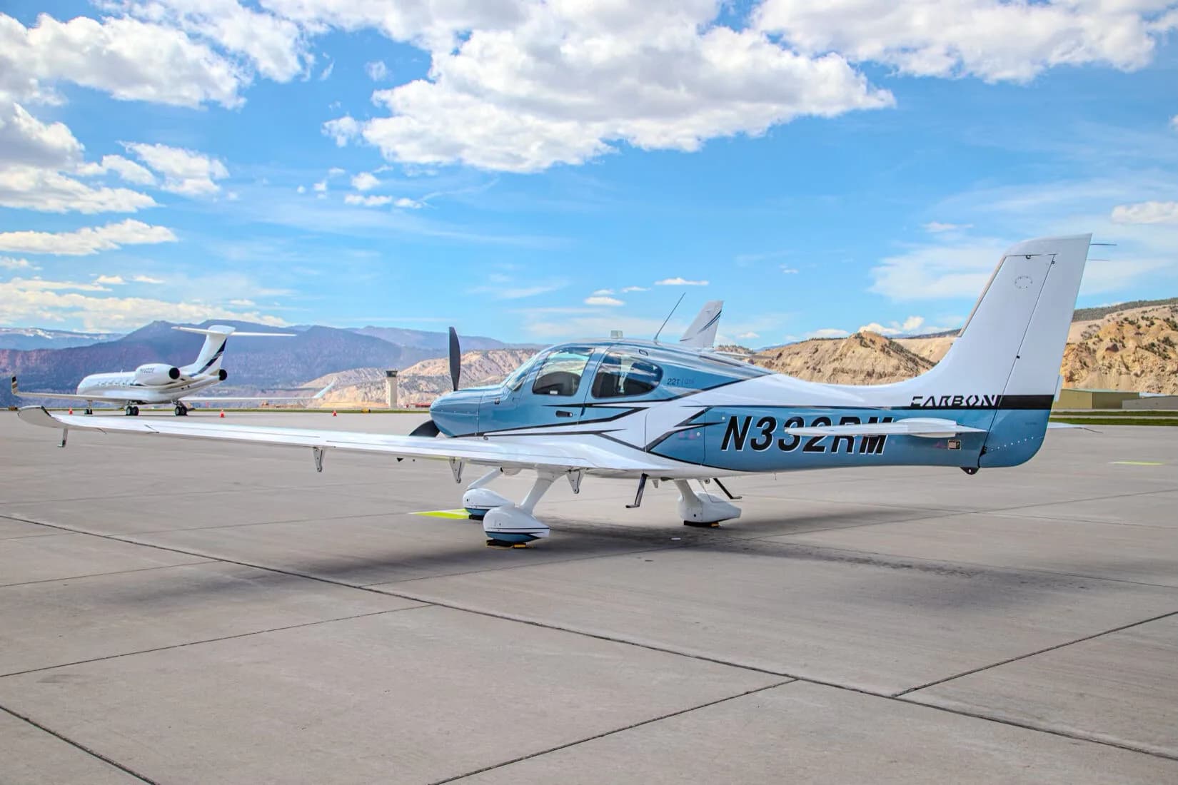 A blue and white Cirrus SR22T GTS aircraft, registration N332RM, parked on an airport tarmac with a private jet in the background.