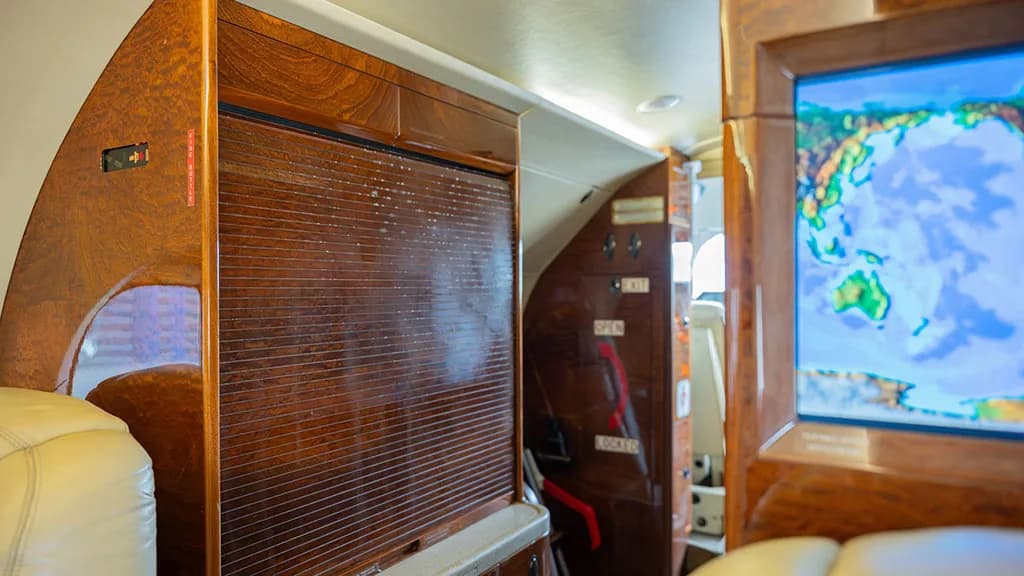 A view inside the private jet cabin showing wood paneling, a closed storage compartment, and a screen displaying a world map.