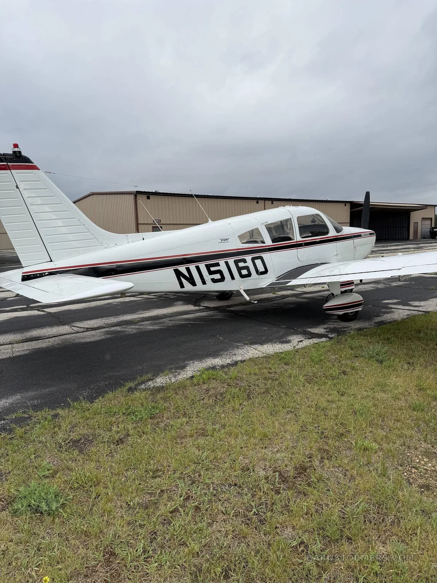 A white Piper Cherokee aircraft with black and red stripes, registration N15160, parked on an asphalt ramp next to grass and hangars.