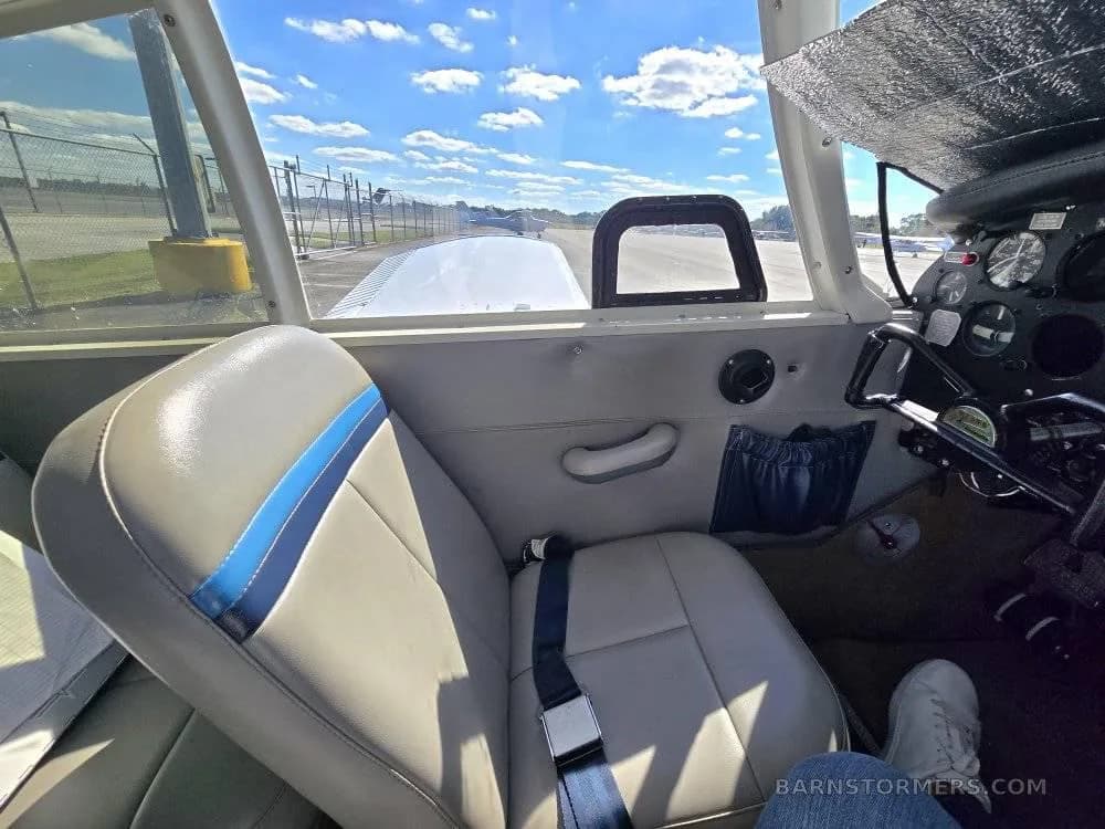 View from inside a light aircraft cabin, showing a grey seat with blue stripes and an airport runway outside.
