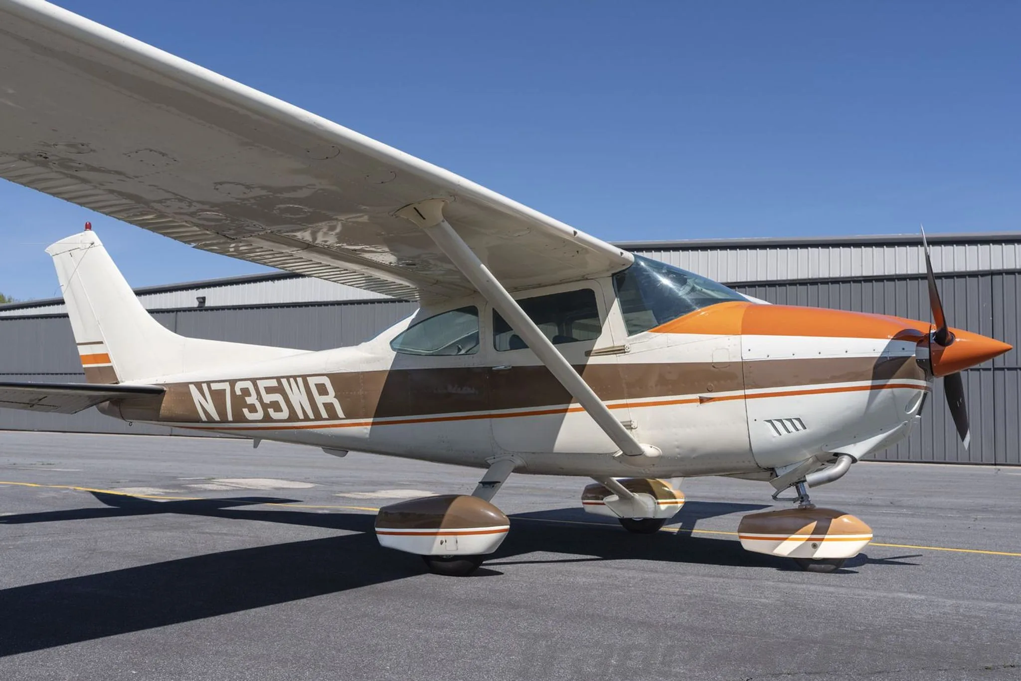 A side view of a white and brown Cessna 172 with orange accents, parked on an asphalt ramp next to a hangar.