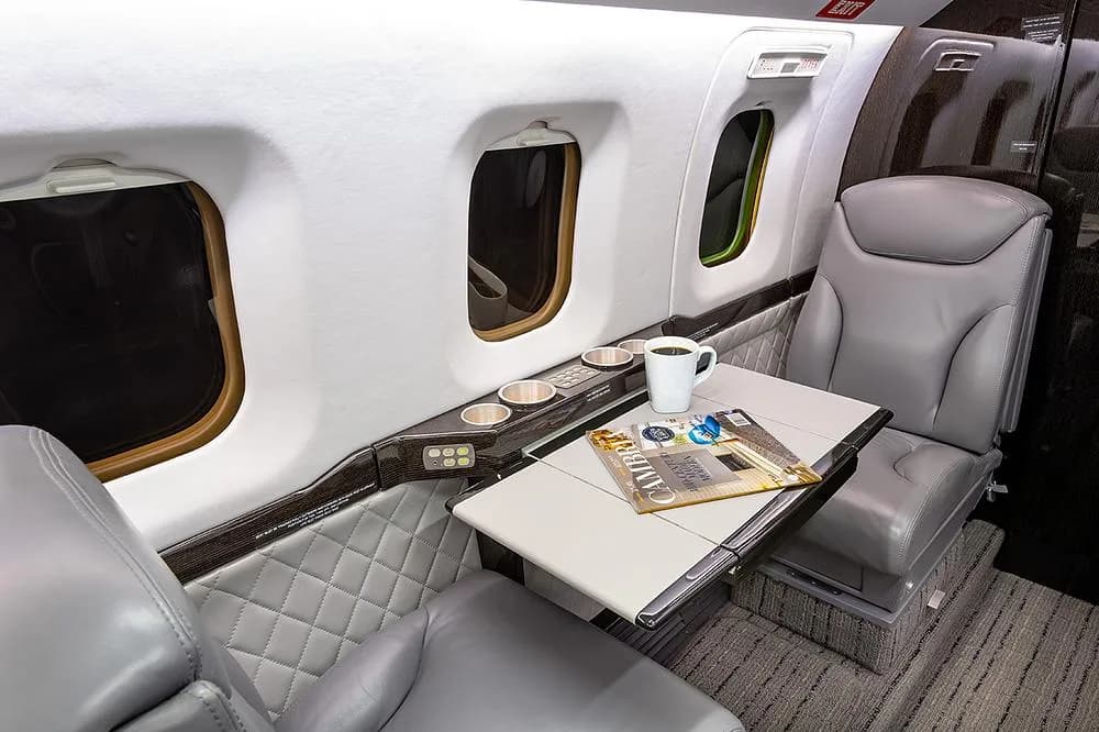 A close-up of the business jet's cabin interior, showing gray leather seats, a fold-out table with a coffee cup, and windows.