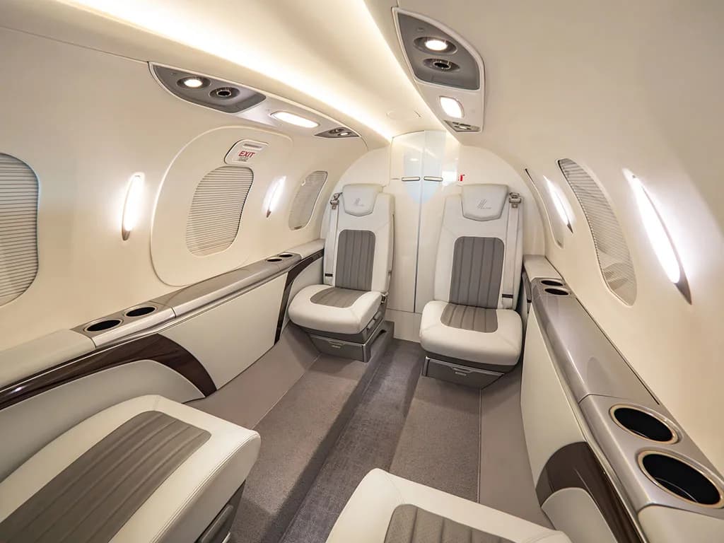 The interior cabin of a HondaJet shows light grey and white leather seats, an aisle, and overhead lighting, looking towards the rear.
