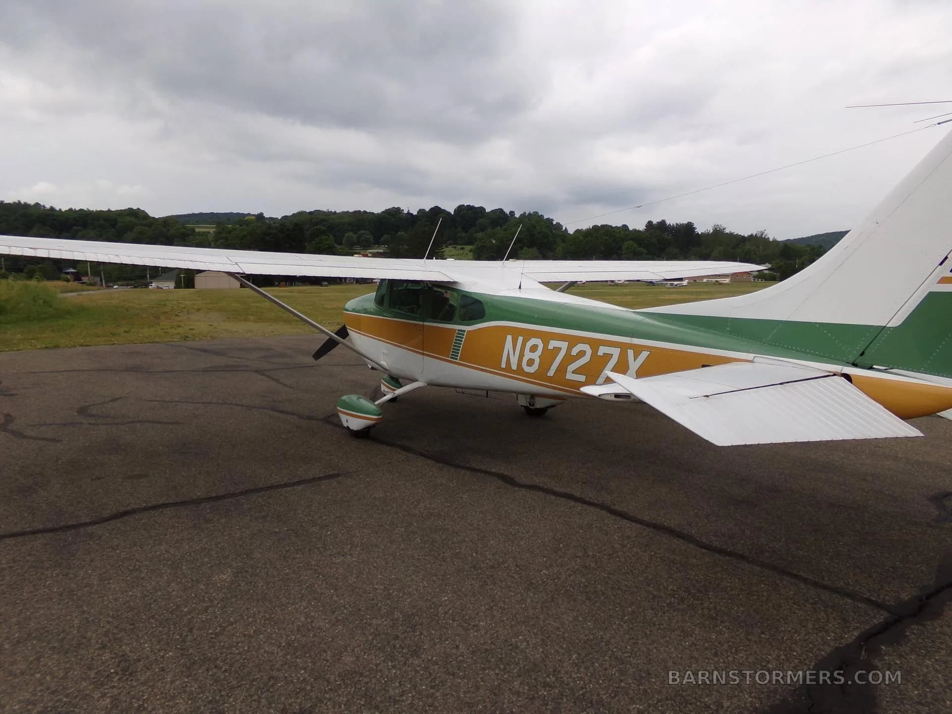 A green, gold, and white Cessna 172 aircraft with registration N8727X, viewed from the rear-left on an asphalt ramp under a cloudy sky.