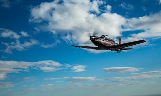 A Beechcraft T-6C Texan II aircraft in flight against a blue sky with clouds.