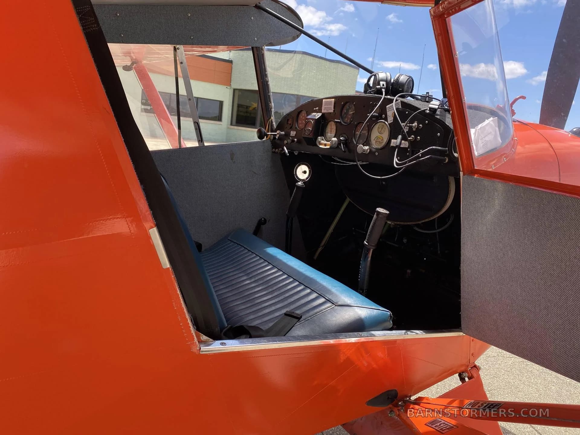 An orange single-engine aircraft with its door open, revealing a blue seat and the instrument panel.